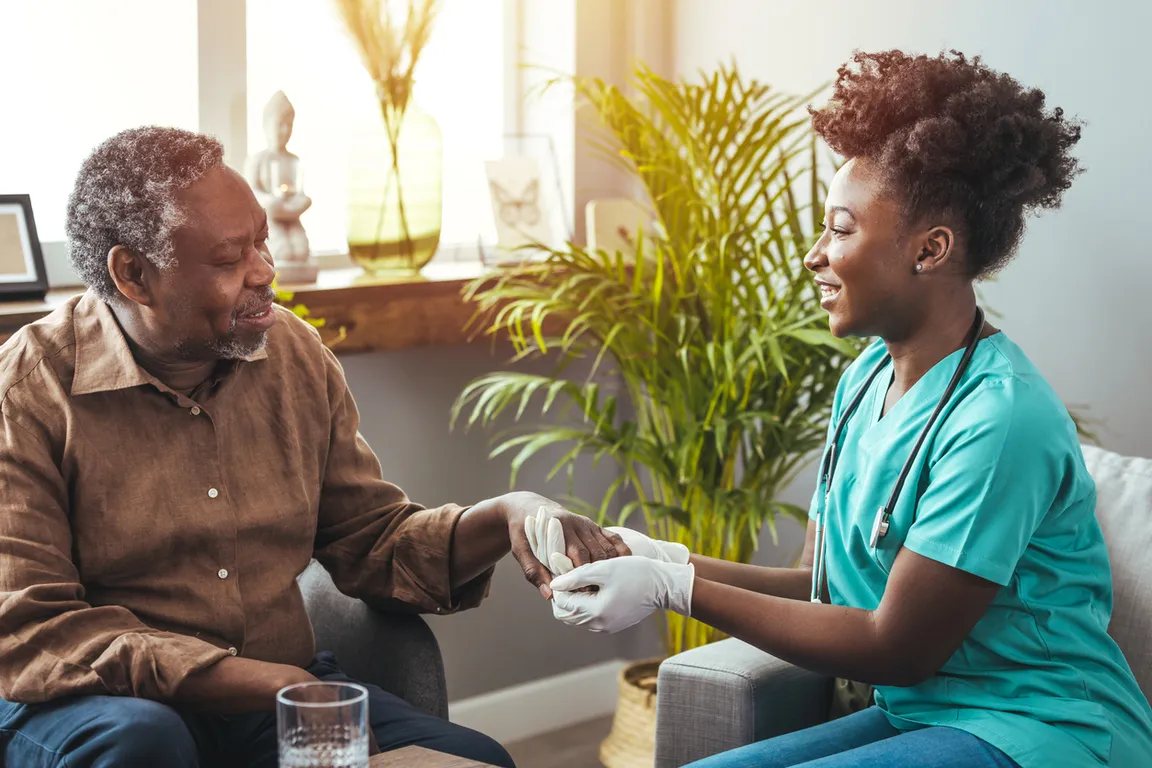 A nurse in scrubs holding an older man’s hand during a home visit